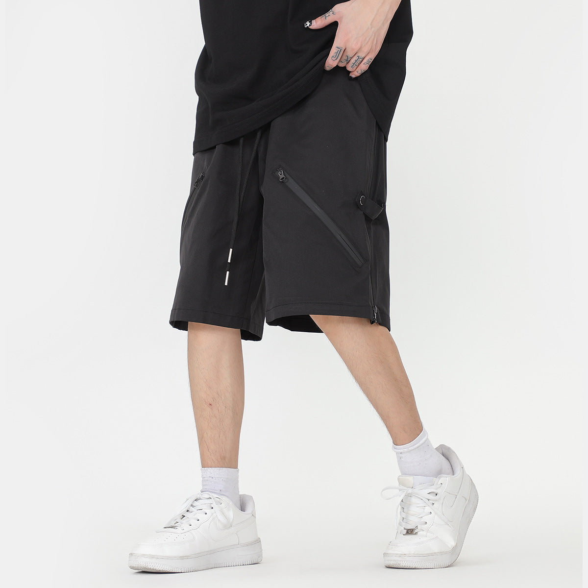 Man wearing black techwear-style cargo shorts with angled zip pockets and white sneakers.