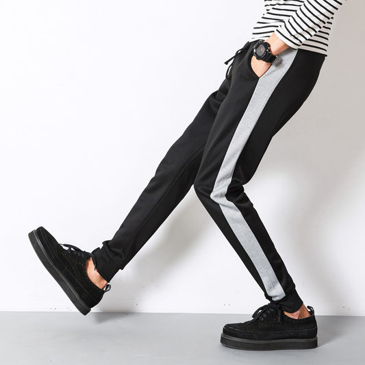 Man wearing black slim fit jogger pants with gray side stripe paired with black sneakers against a light wall 
background. 