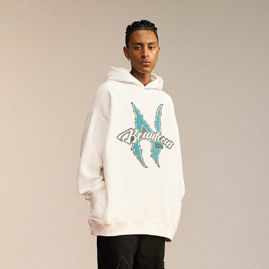 Man wearing an oversized cream streetwear hoodie with a bold “Beautcous” logo graphic and teal claw-style 
design on the front. 