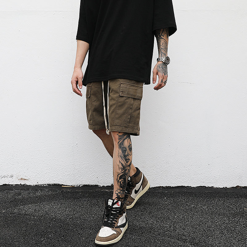 Man wearing relaxed fit brown cargo shorts with oversized black t-shirt and sneakers in a streetwear outfit 
against a white wall. 
