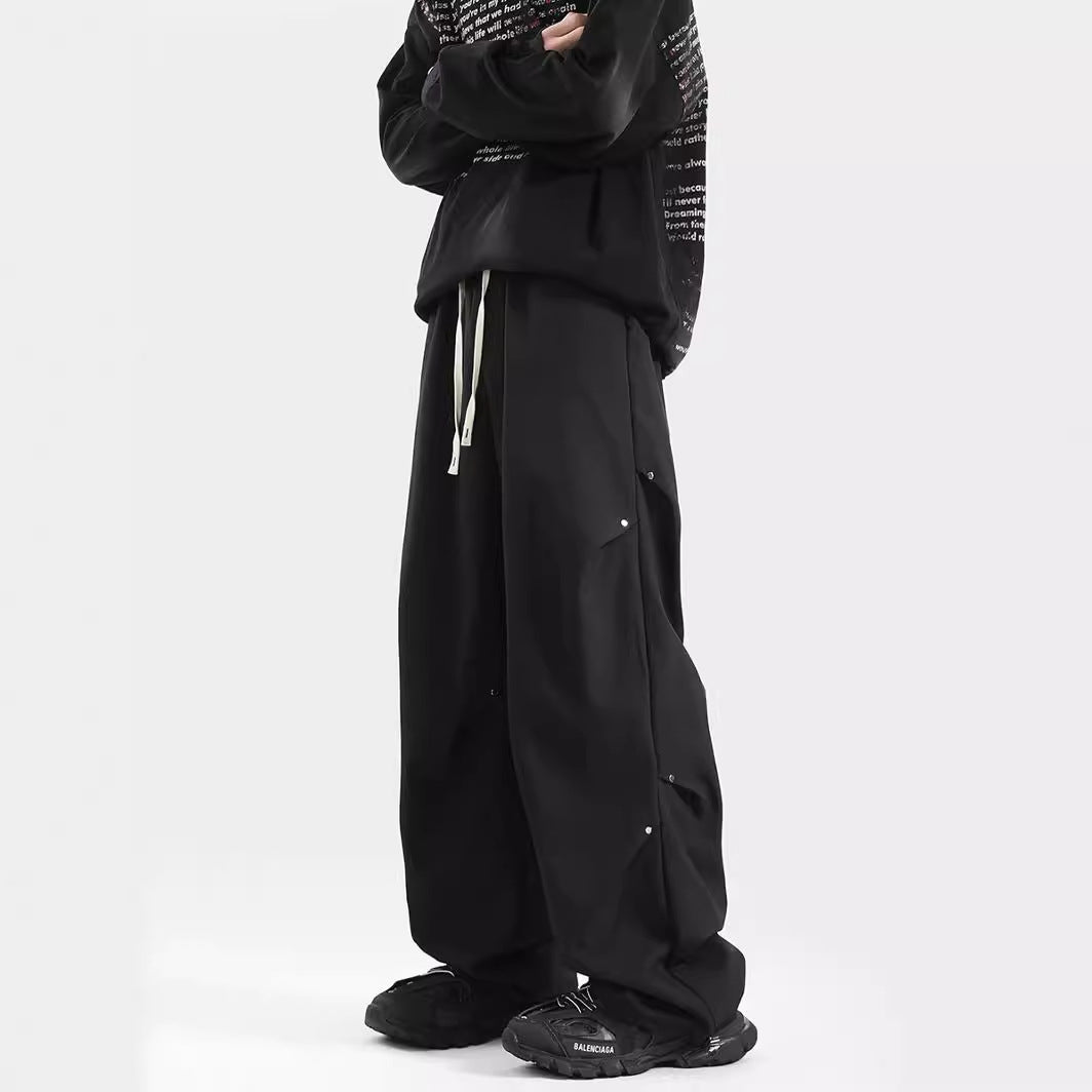 Men’s black oversized techwear cargo pants with drawstring waistband, metallic detail and wide-leg fit worn 
with sneakers against a white background. 
