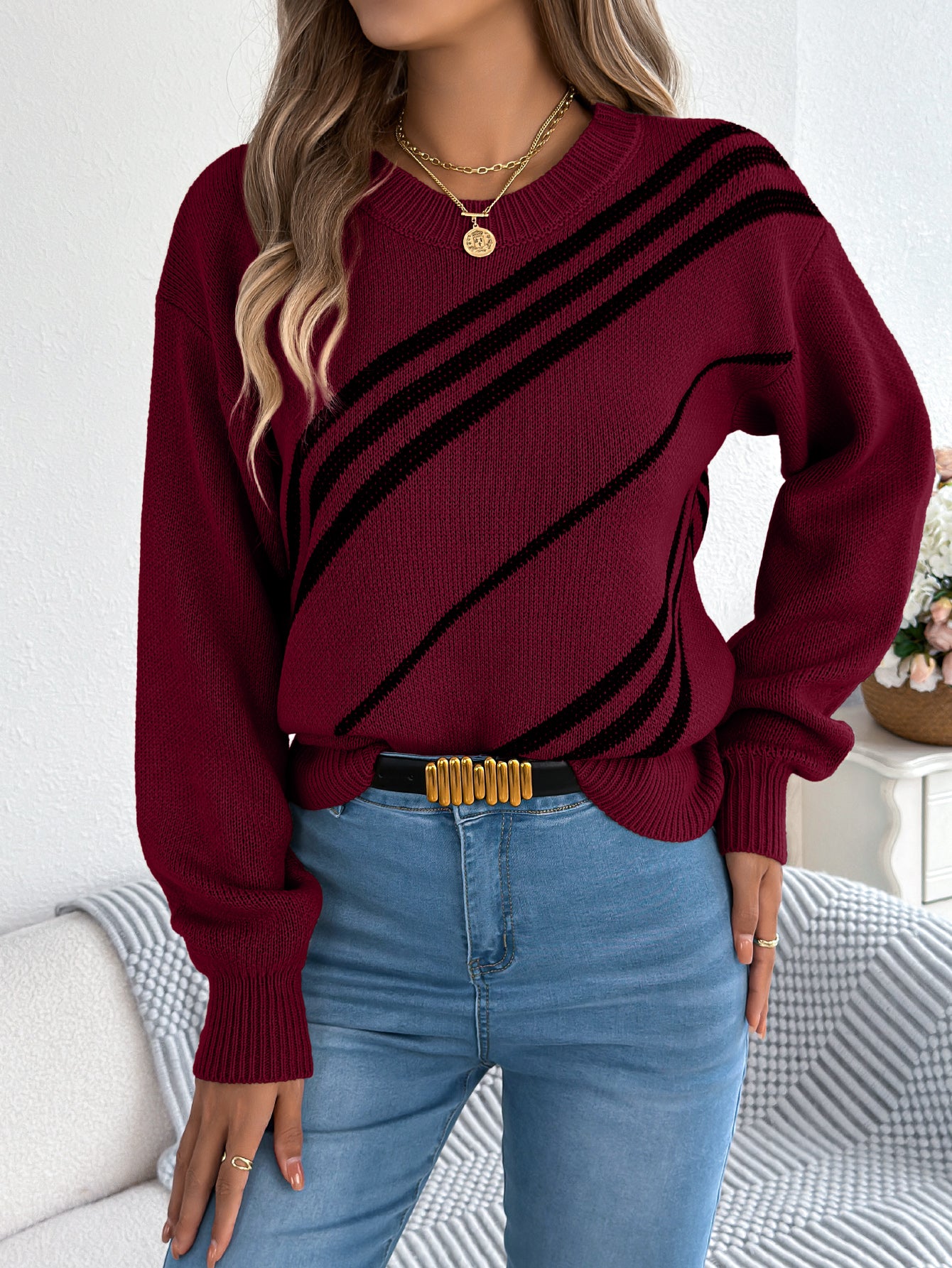 Woman wearing amaroon knit sweater with black diagonal stripes, styled with blue jeans and gold necklace. 