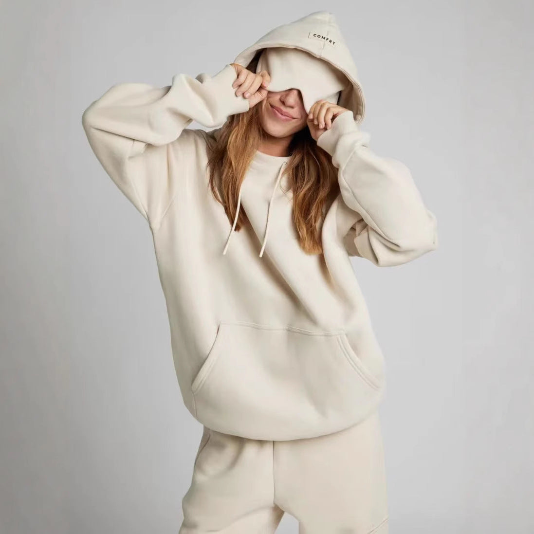 Woman wearing an oversized cream fleece hoodie with eye-mask hood detail, drawstring hood, and front 
pouch pocket. 