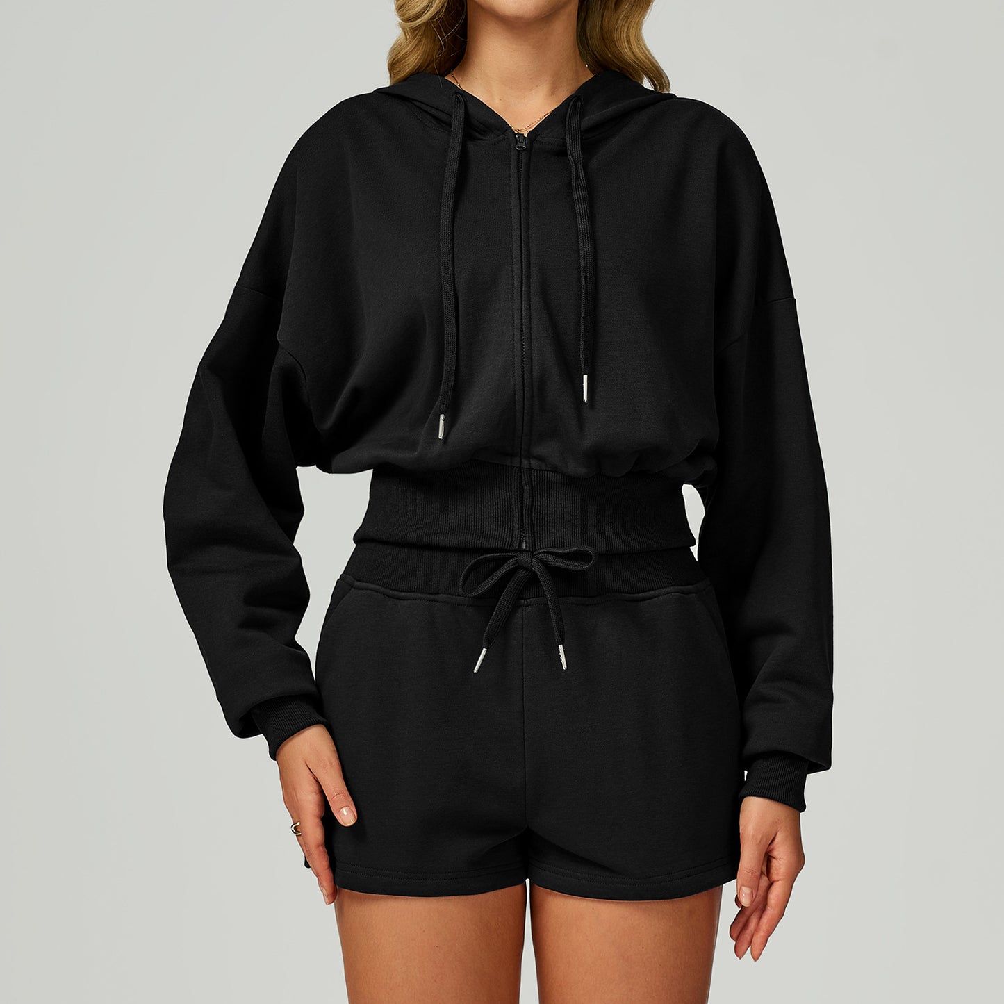 Woman wearing a black cropped zip-up hoodie with drawstring hood and shorts set. 